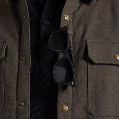 Close-up of a person wearing an olive brown textured jacket over a black knit shirt, with black sunglasses resting on the chest pocket.