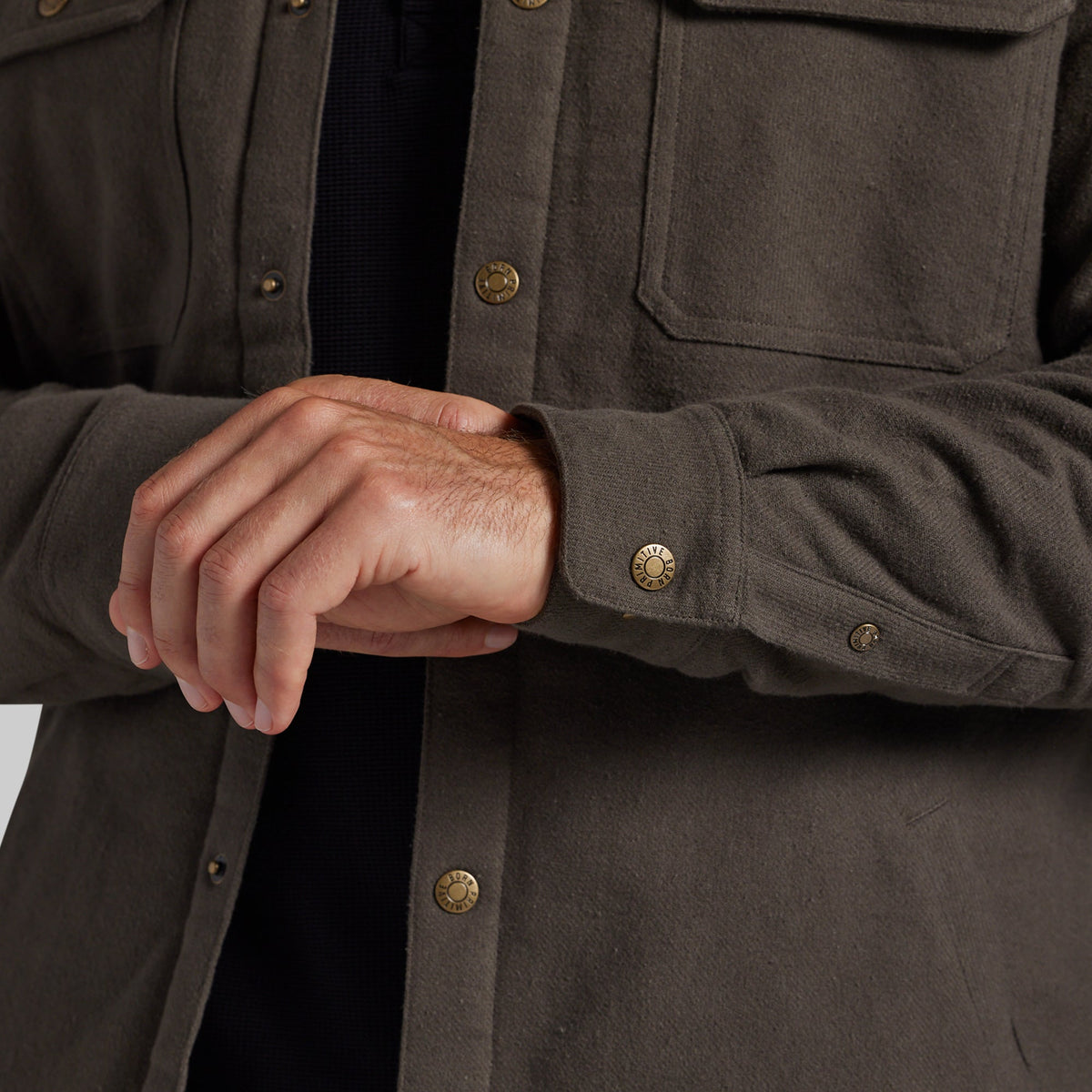 Close-up of a person wearing a dark olive-brown overshirt with brass buttons over a black shirt, showing the hands adjusting or fastening the cuff.