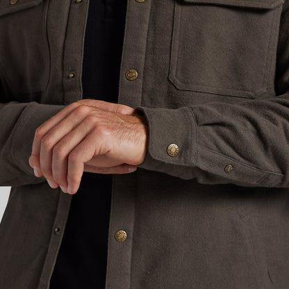 Close-up of a person wearing a dark olive-brown overshirt with brass buttons over a black shirt, showing the hands adjusting or fastening the cuff.