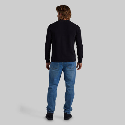A man viewed from behind wearing a black long-sleeve crewneck sweater, blue jeans, and black shoes, standing against a solid light gray background.