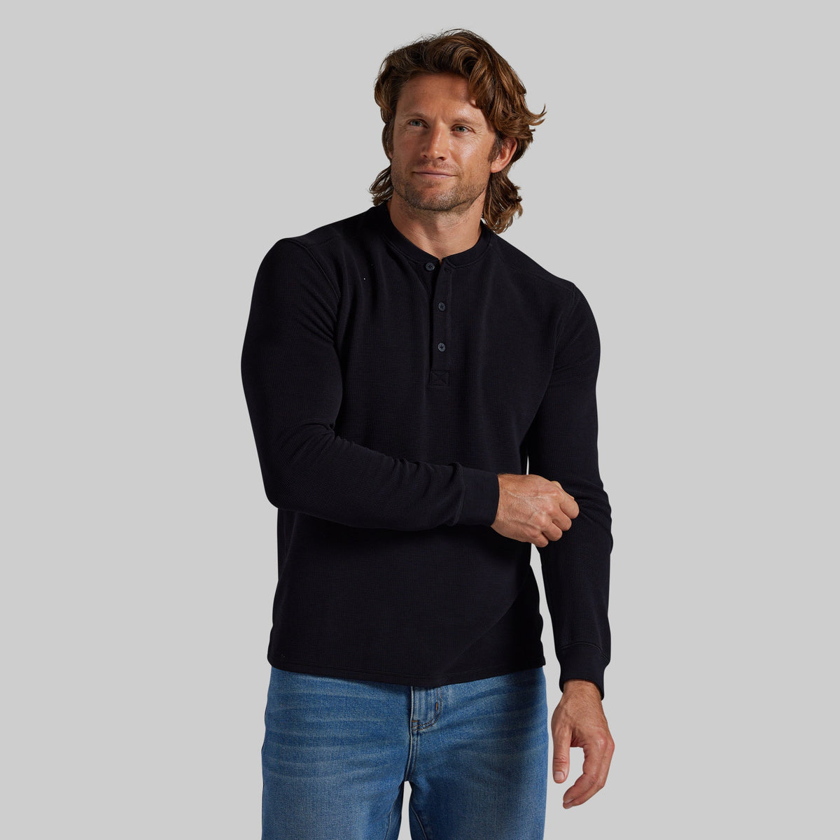 A man wearing a black long-sleeve henley shirt and blue jeans, standing against a solid gray studio background.