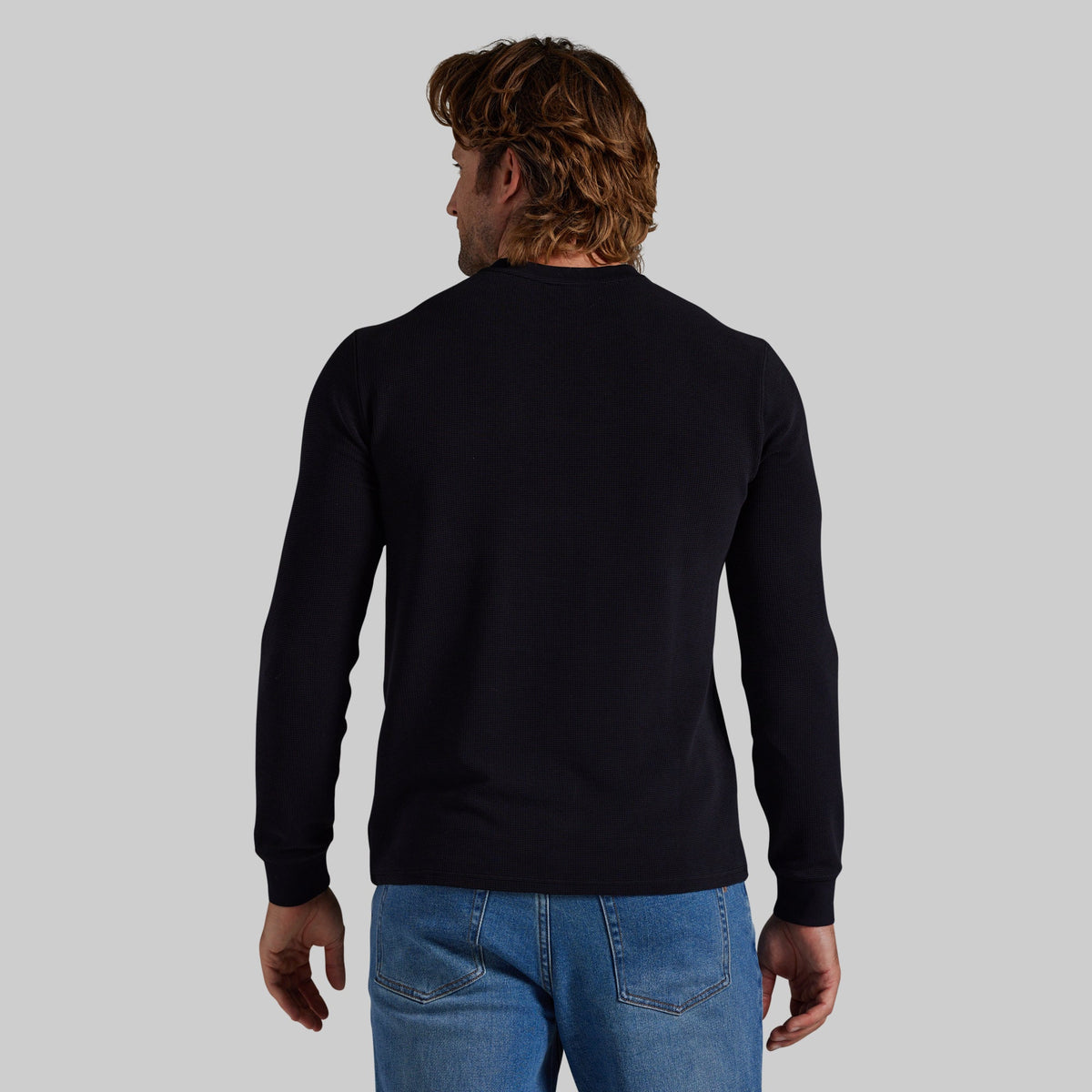 A man viewed from behind wearing a black long-sleeve crewneck sweater and blue jeans, set against a solid light gray background.