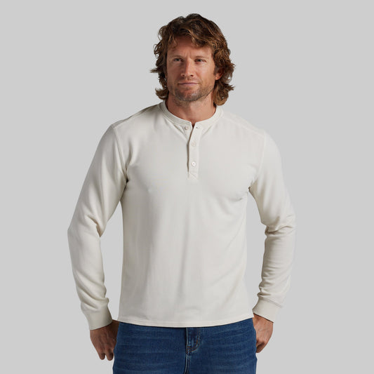 A man wearing a cream-colored long-sleeve henley shirt with blue jeans, standing against a solid gray studio background.