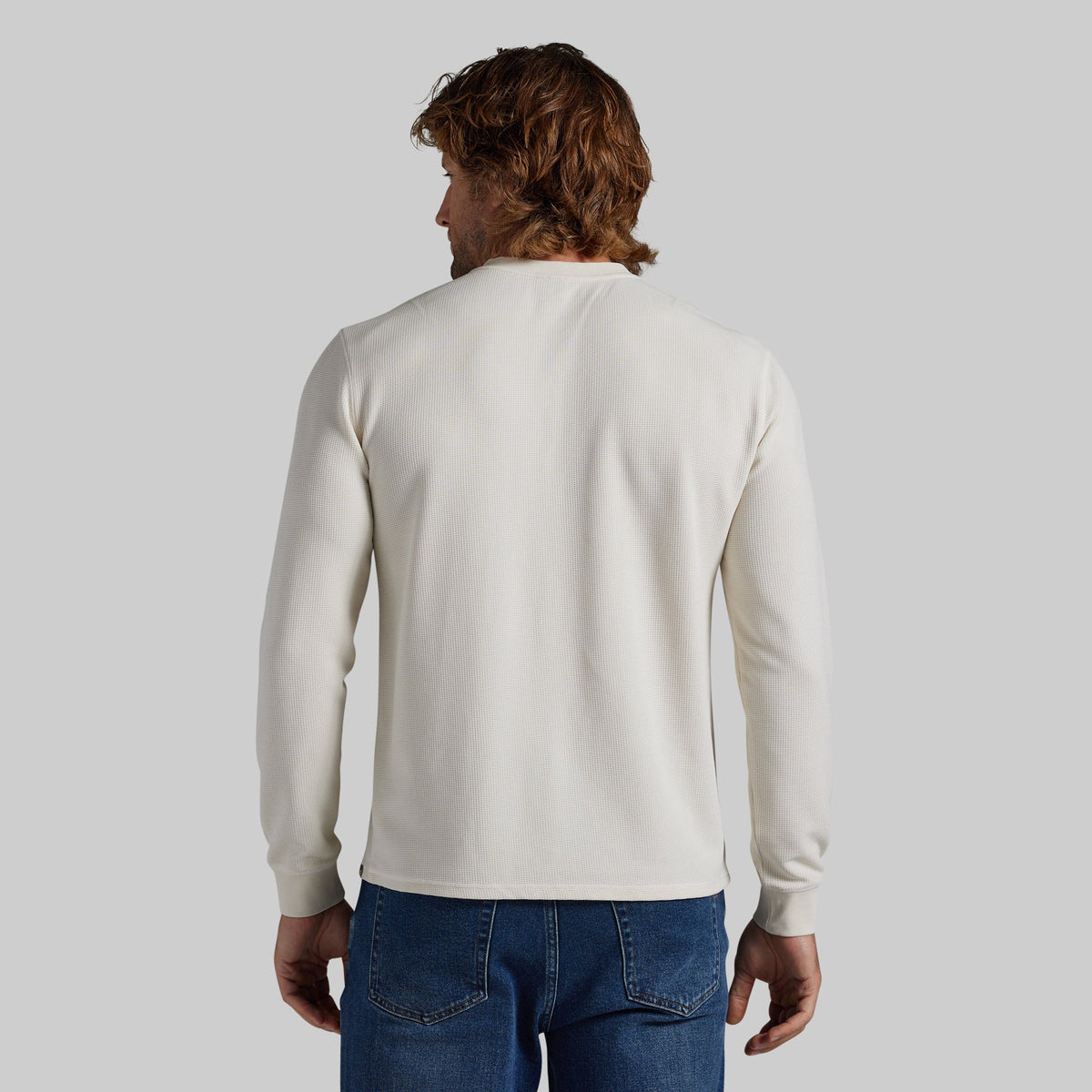 A man viewed from behind wearing a plain off-white long-sleeve crewneck sweater and blue jeans, set against a solid light gray background.