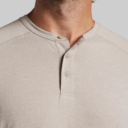 Close-up of a man wearing a light beige heather henley shirt, showing the collar, three-button placket, and chest fabric against a plain light gray background.