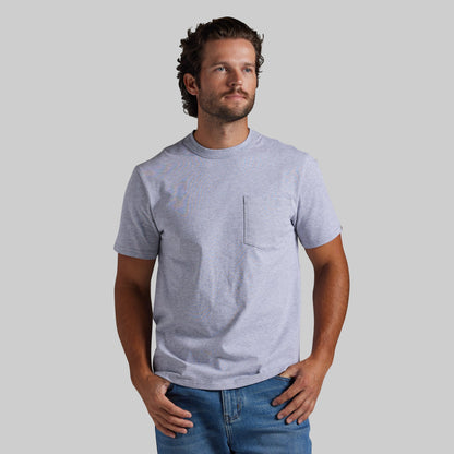 A man wearing a light heather gray short-sleeve crewneck t-shirt with a chest pocket, paired with blue jeans, standing against a solid gray background.