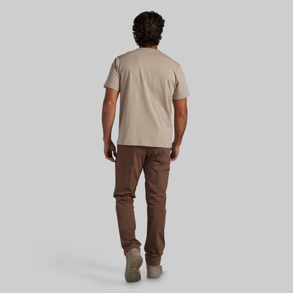 A man shown from behind wearing a beige short-sleeve t-shirt, brown pants, and beige shoes, walking against a solid light gray background.