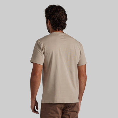 A man viewed from behind wearing a light beige short-sleeve t-shirt, paired with brown pants, against a solid gray background.