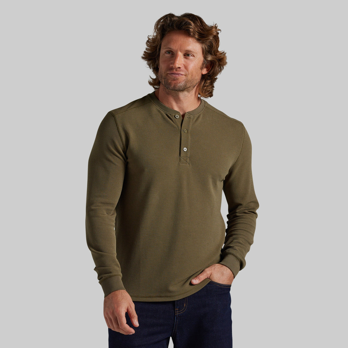 A man wearing a long-sleeve henley shirt in olive green, paired with dark blue jeans, standing against a solid gray background.