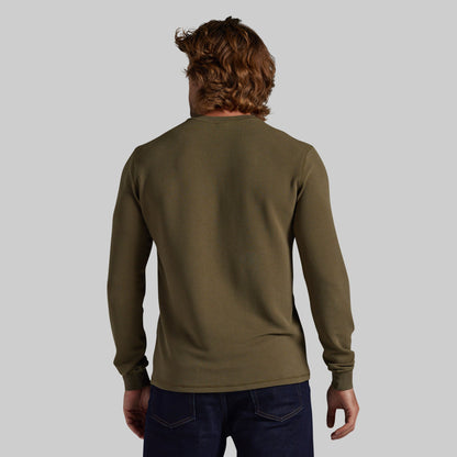 A man viewed from behind wearing an olive green long-sleeve shirt and dark blue jeans, set against a solid light gray background.