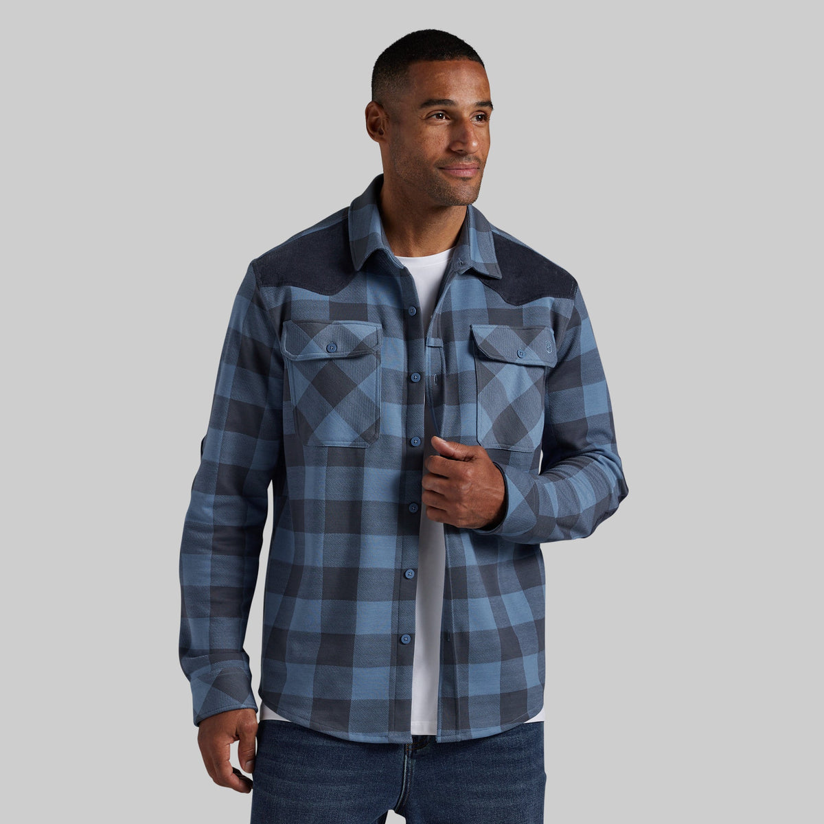 A man wearing a blue and gray plaid button-up shirt with dark navy shoulder panels, layered over a white t-shirt and blue jeans, set against a solid gray background.