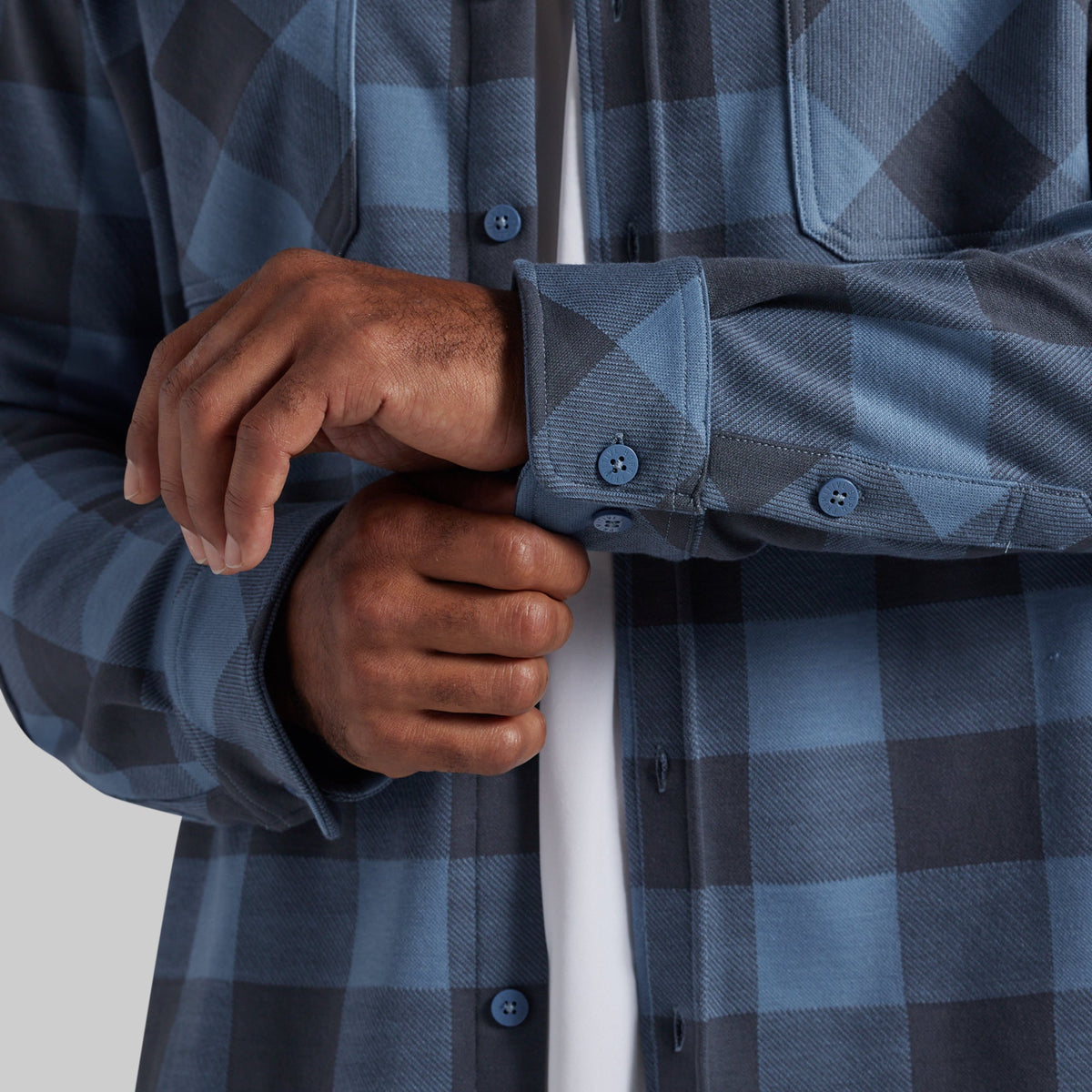 Close-up of a person wearing a blue and gray buffalo plaid flannel shirt, adjusting the cuff of the right sleeve over a white undershirt.