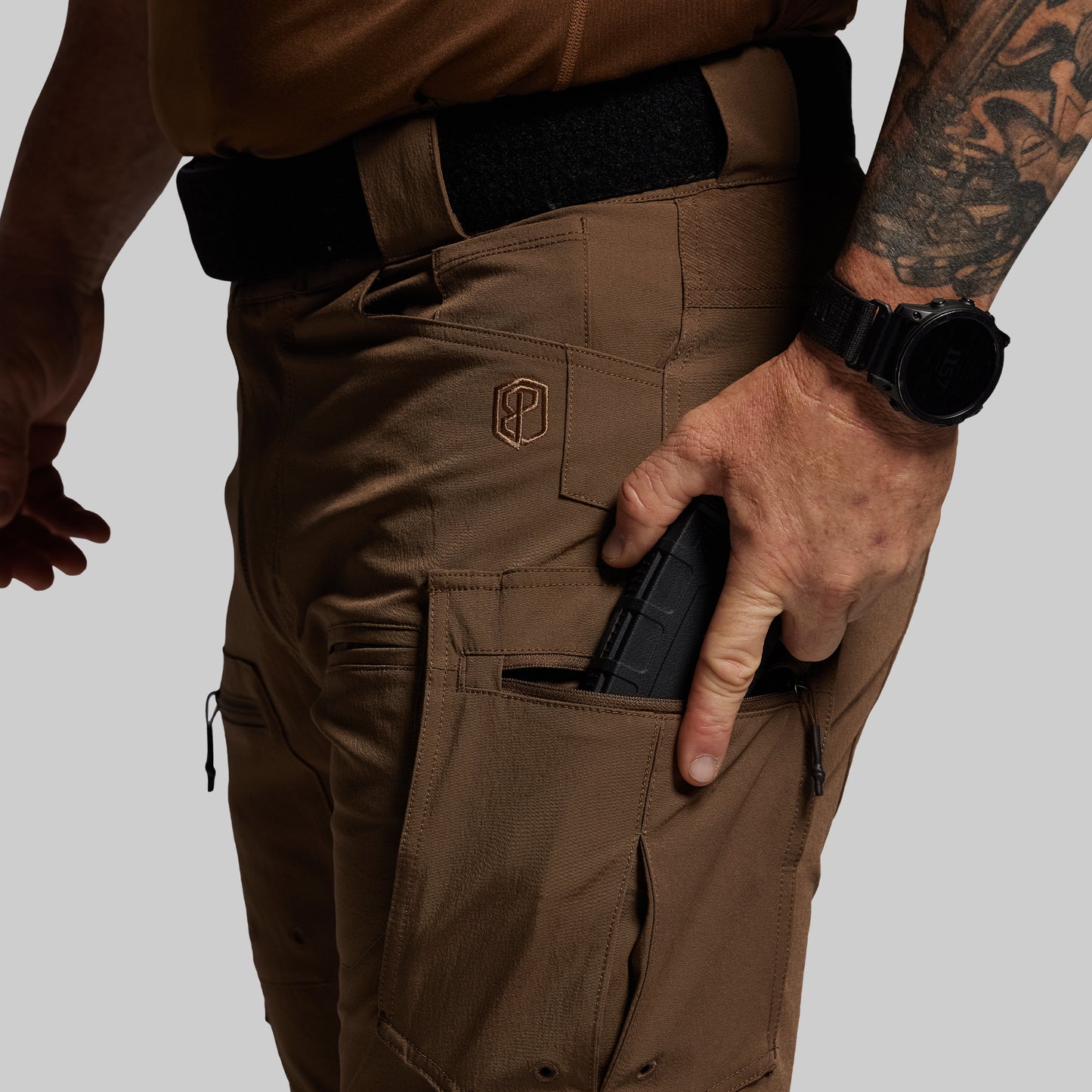 Person wearing brown tactical pants with a visible brand logo, holding a magazine.