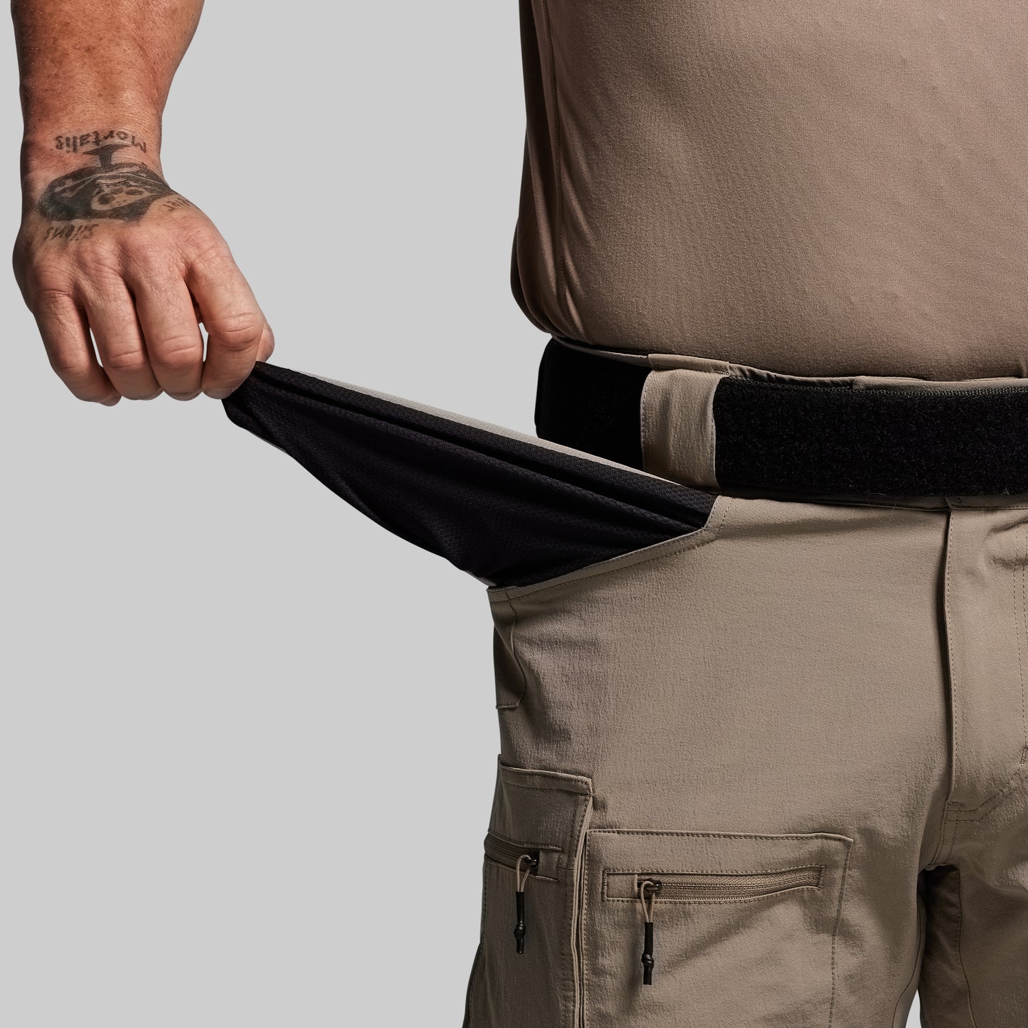 Person wearing tan pants with a black belt, showing the inside of front pocket on a gray background 