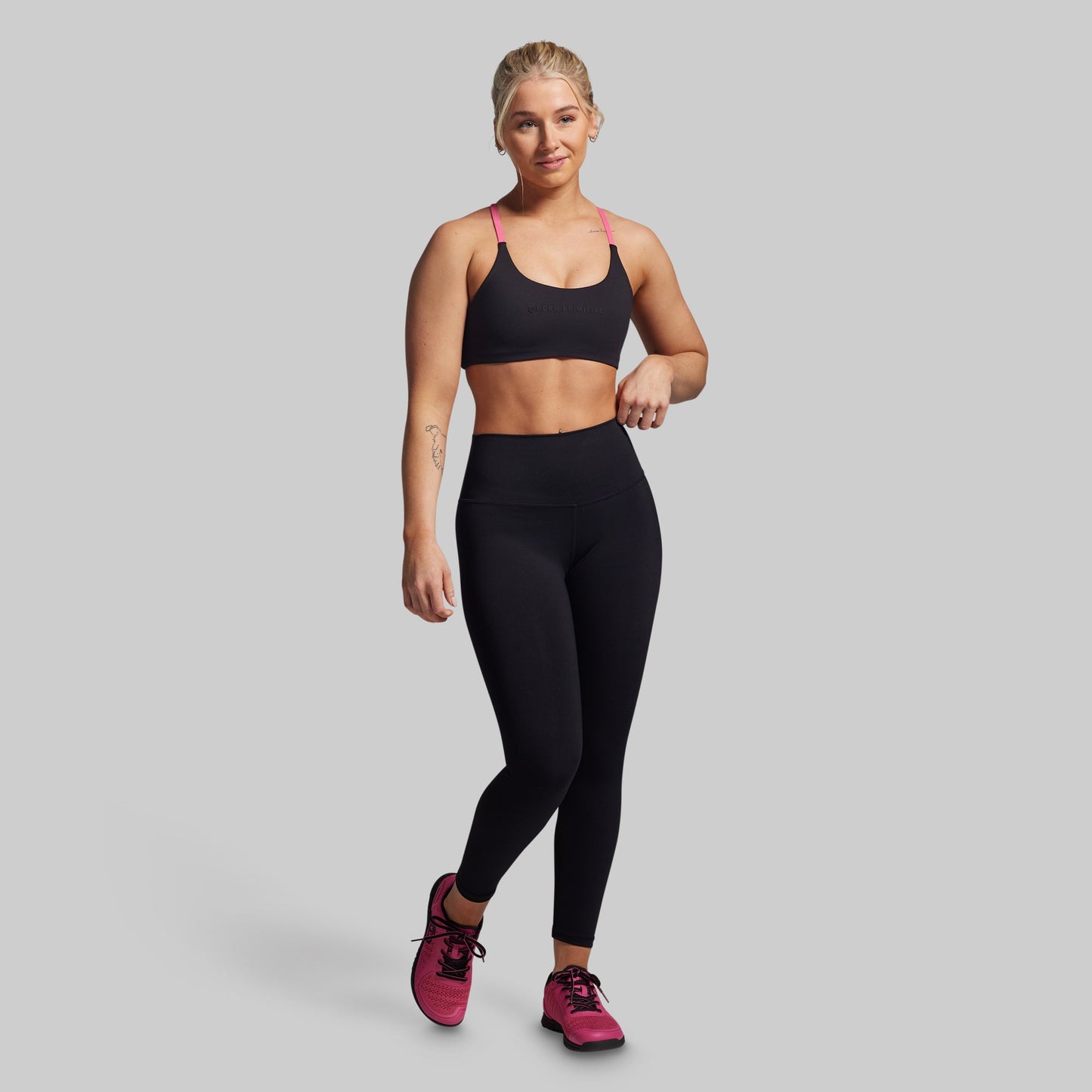 Woman wearing a black sports bra and leggings with pink shoes on a gray background