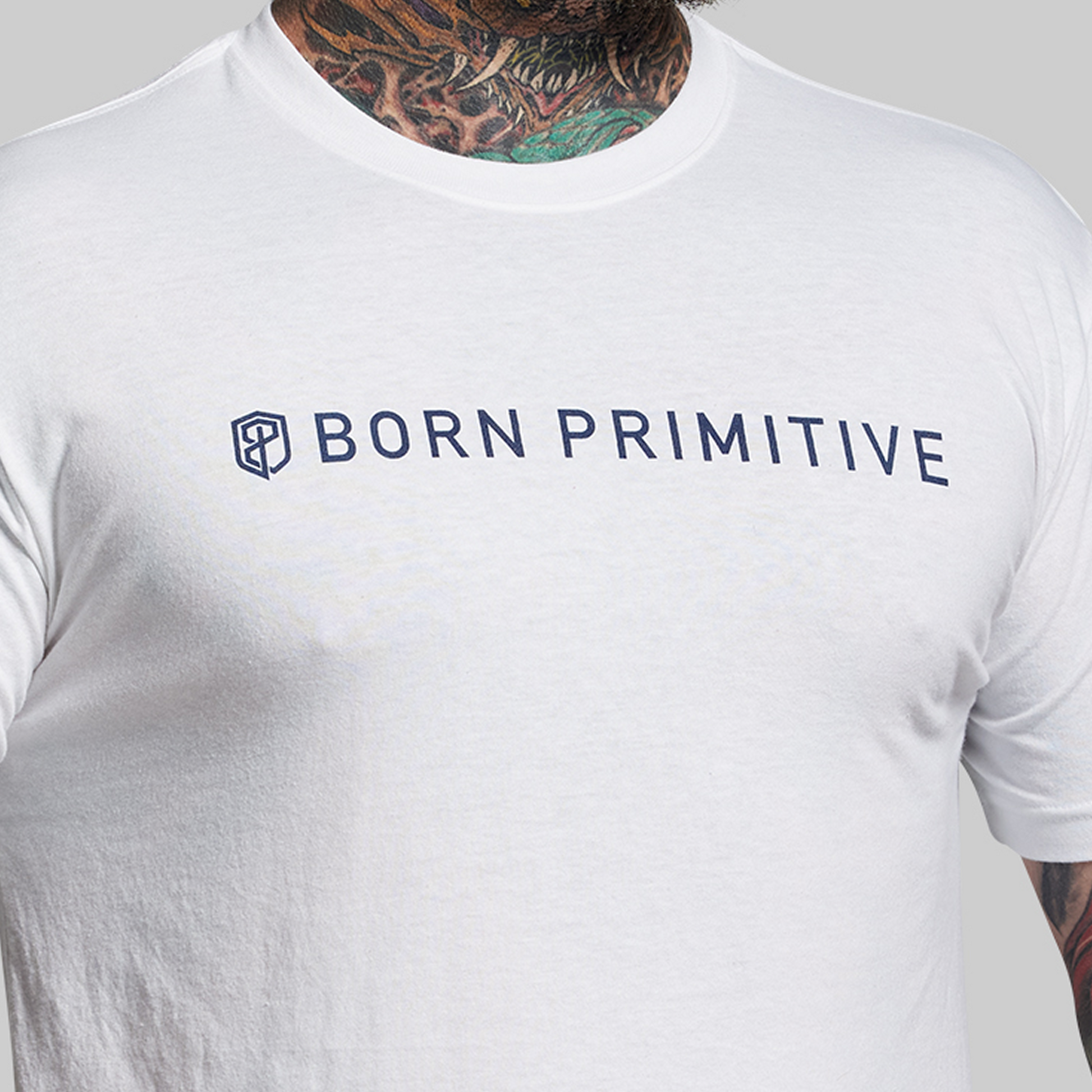 White t-shirt with 'BORN PRIMITIVE' text worn by a person with tattoos on a gray background