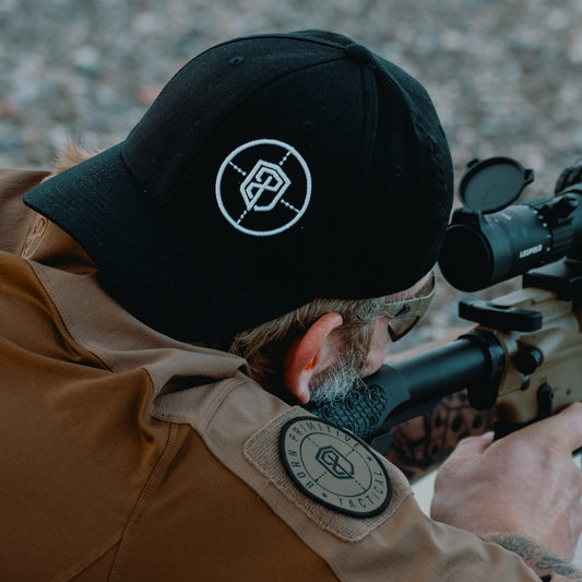 Person wearing a black cap and tan long sleeve with a logo, looking through scope.