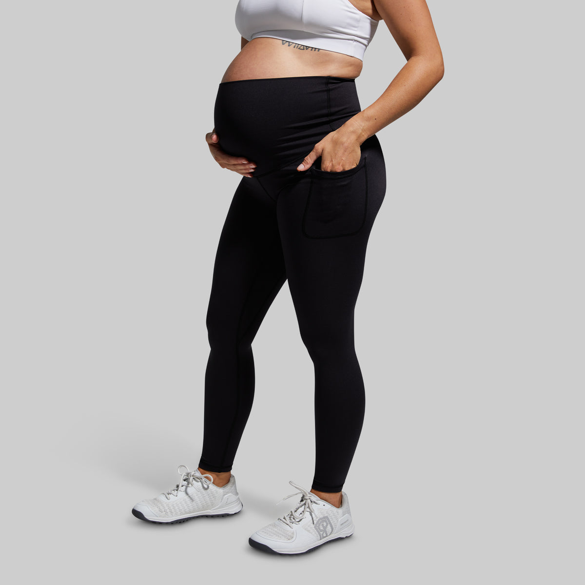A pregnant model wearing black high-waisted maternity leggings with a side pocket, paired with a white sports bra and white sneakers, shown from the waist down against a solid gray background.