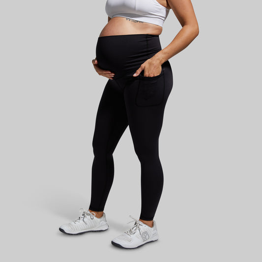 A pregnant model wearing black high-waisted maternity leggings with a side pocket, paired with a white sports bra and white sneakers, shown from the waist down against a solid gray background.