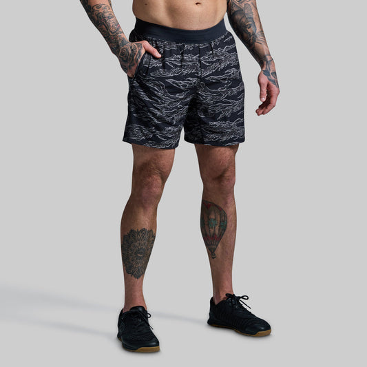 A person wearing a camouflage patterned compression short with a quick-dry outer short over it. The person is standing with hands in pockets and wearing black sneakers.
