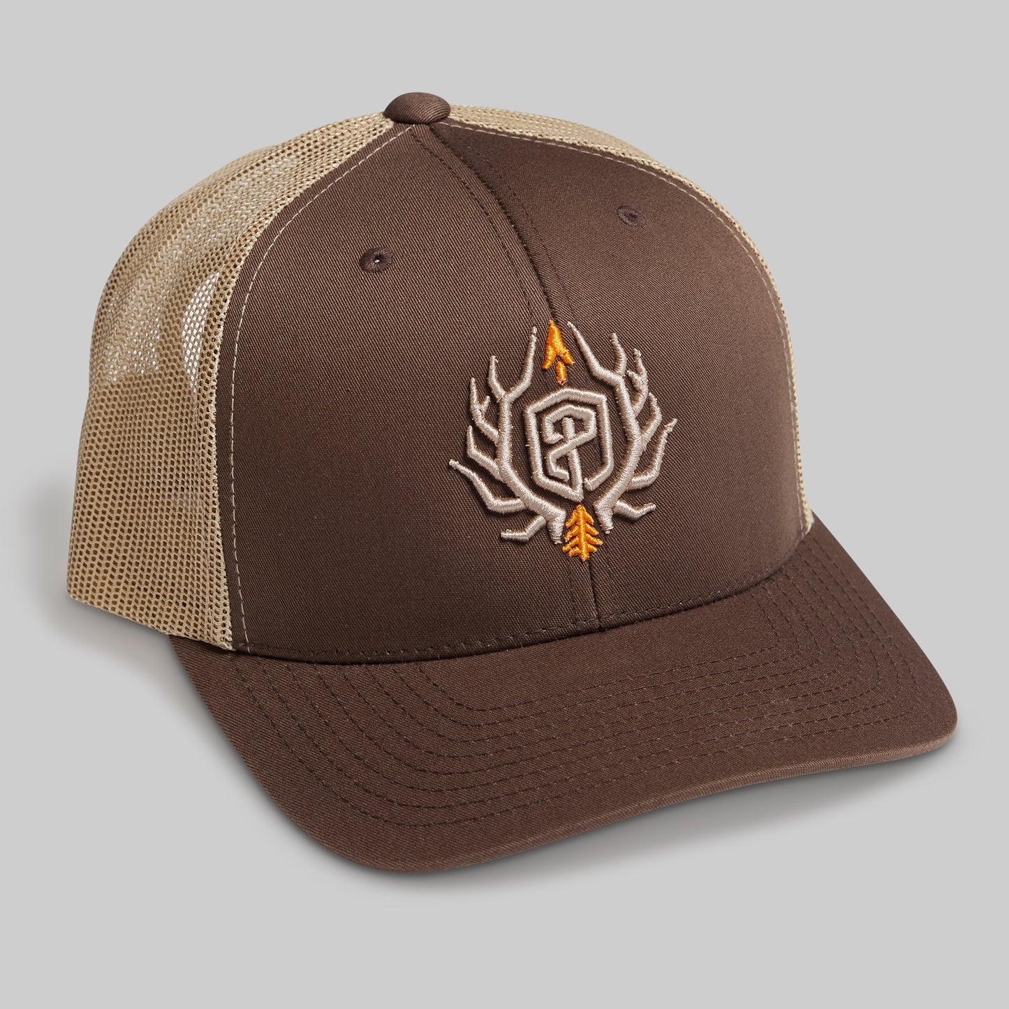 A brown and tan trucker-style cap with a brown front panel and brim, tan mesh back, and an embroidered antlered monogram logo on the front in brown, white, and orange.