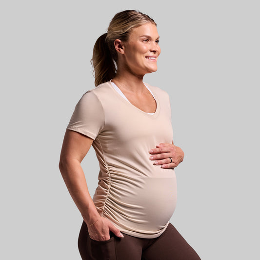 A pregnant woman wearing a short sleeve beige maternity top with a V-neck, the shirt has ruched sides.