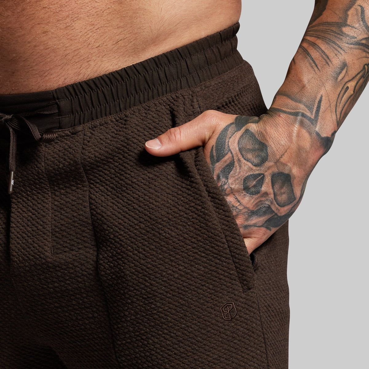 Brown textured joggers worn by a person with a tattooed arm on a gray background