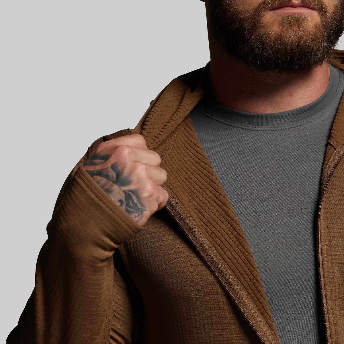 Man wearing a brown hoodie over a gray shirt with a tattoo on his arm, against a plain background