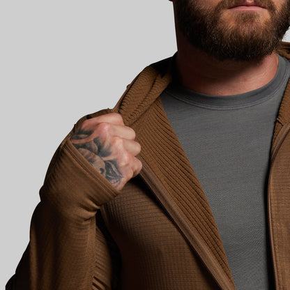 Man wearing a brown hoodie over a gray shirt with a tattoo on his arm, against a plain background