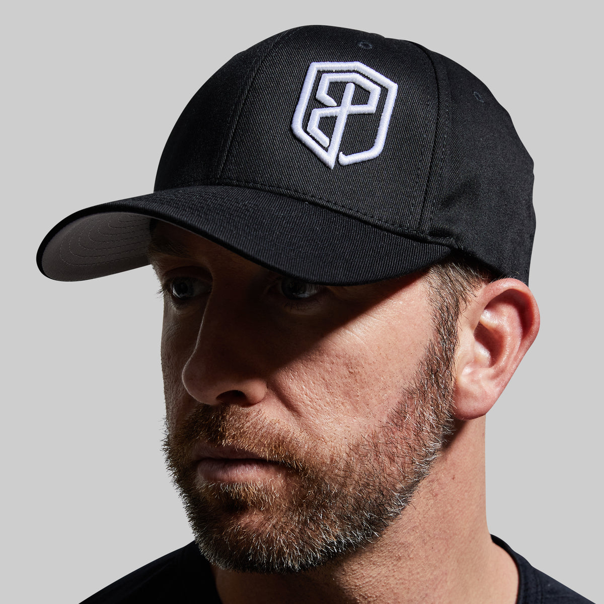 A close-up of a man wearing a black baseball cap with a white embroidered geometric logo on the front, set against a solid light gray background.