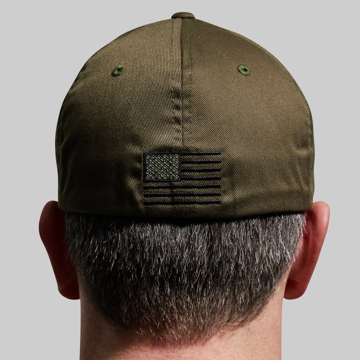 Back view of a man wearing an olive green baseball cap with a black embroidered American flag patch on the back, set against a solid gray background.