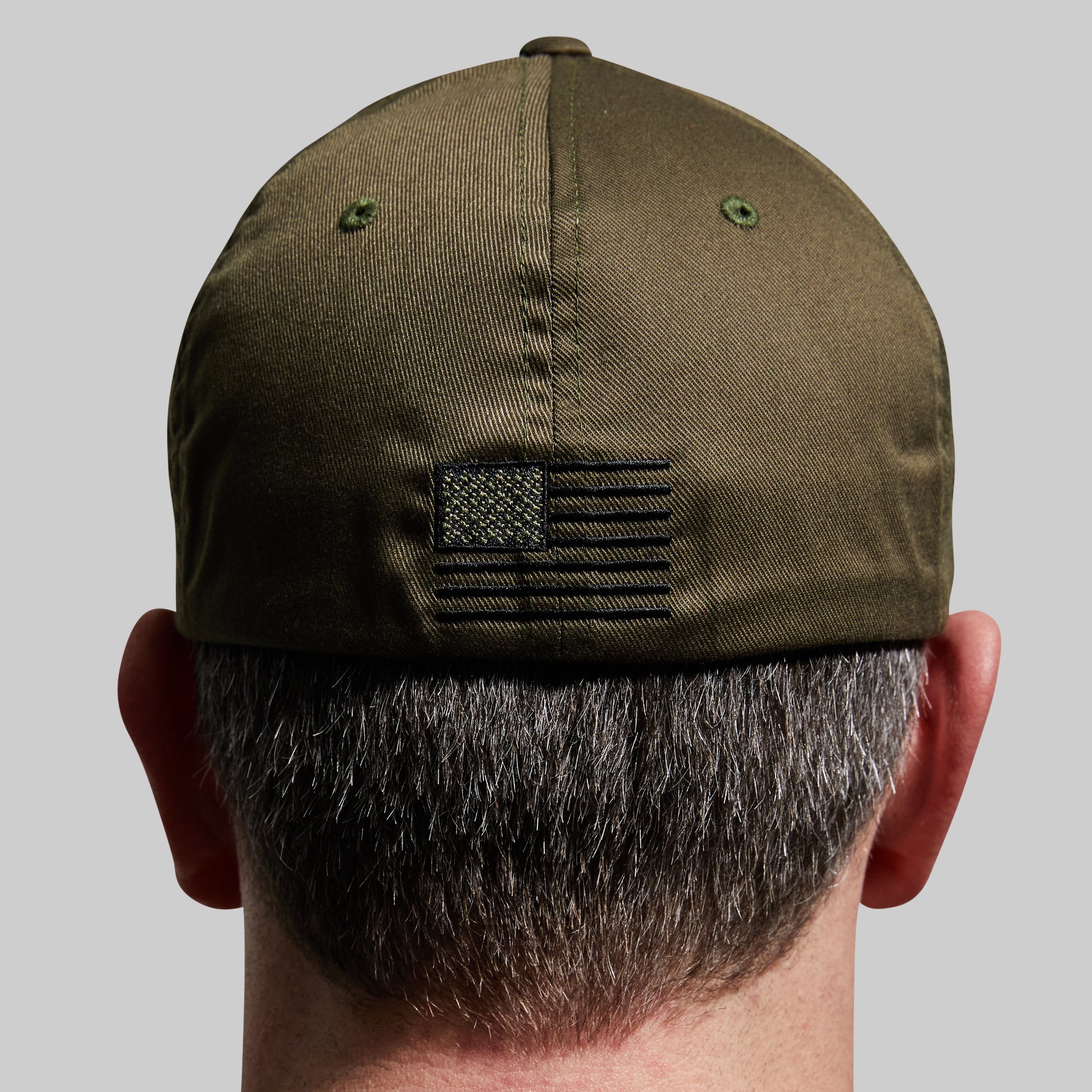 Back view of a man wearing an olive green baseball cap with a black embroidered American flag patch on the back, set against a solid gray background.