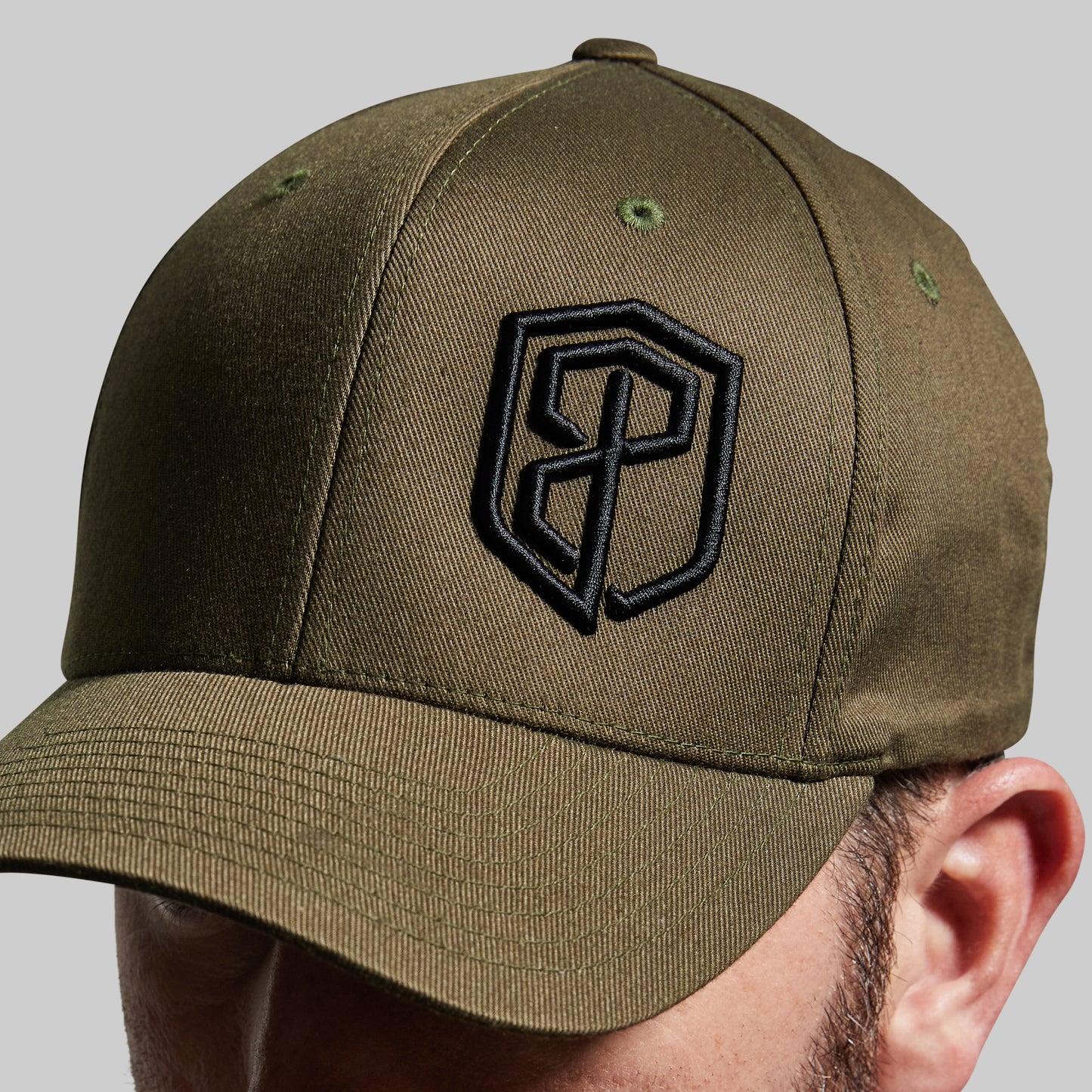 An olive green baseball cap with a black embroidered shield logo on the front, shown worn by a model against a neutral gray background.