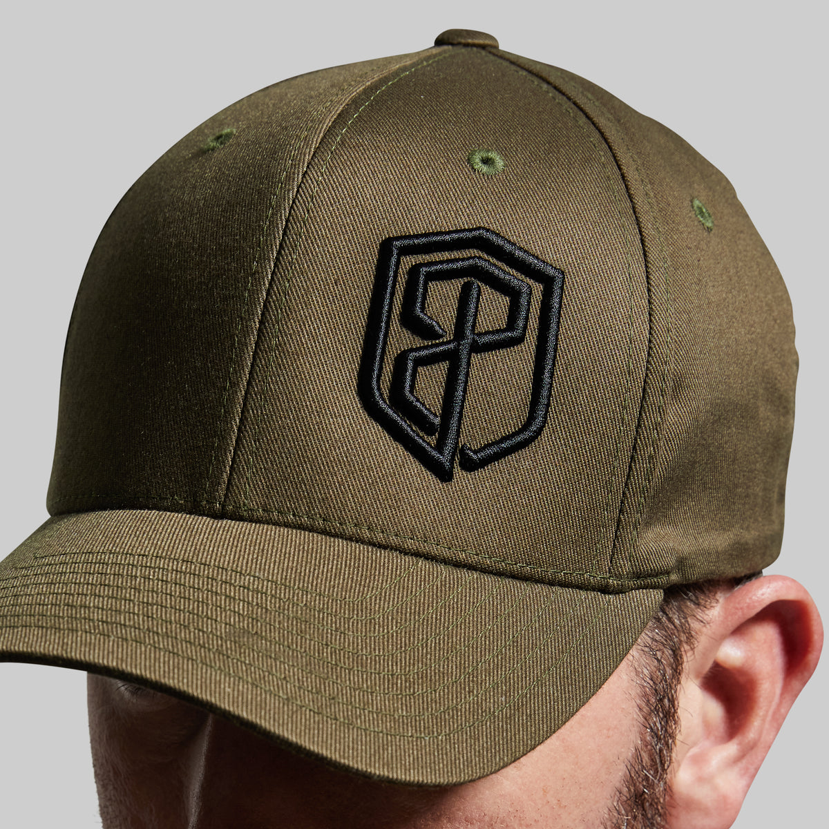 An olive green baseball cap with a black embroidered shield logo on the front, shown worn by a model against a neutral gray background.