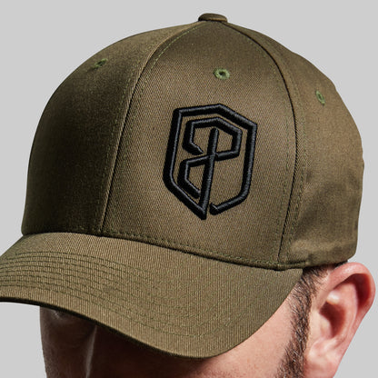 An olive green baseball cap with a black embroidered shield logo on the front, shown worn by a model against a neutral gray background.
