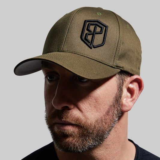 A close-up of a man wearing an olive green baseball cap with a black embroidered shield logo on the front, set against a solid gray background.