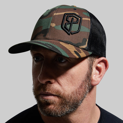 A person wearing a camouflage trucker hat with a black logo on the front.