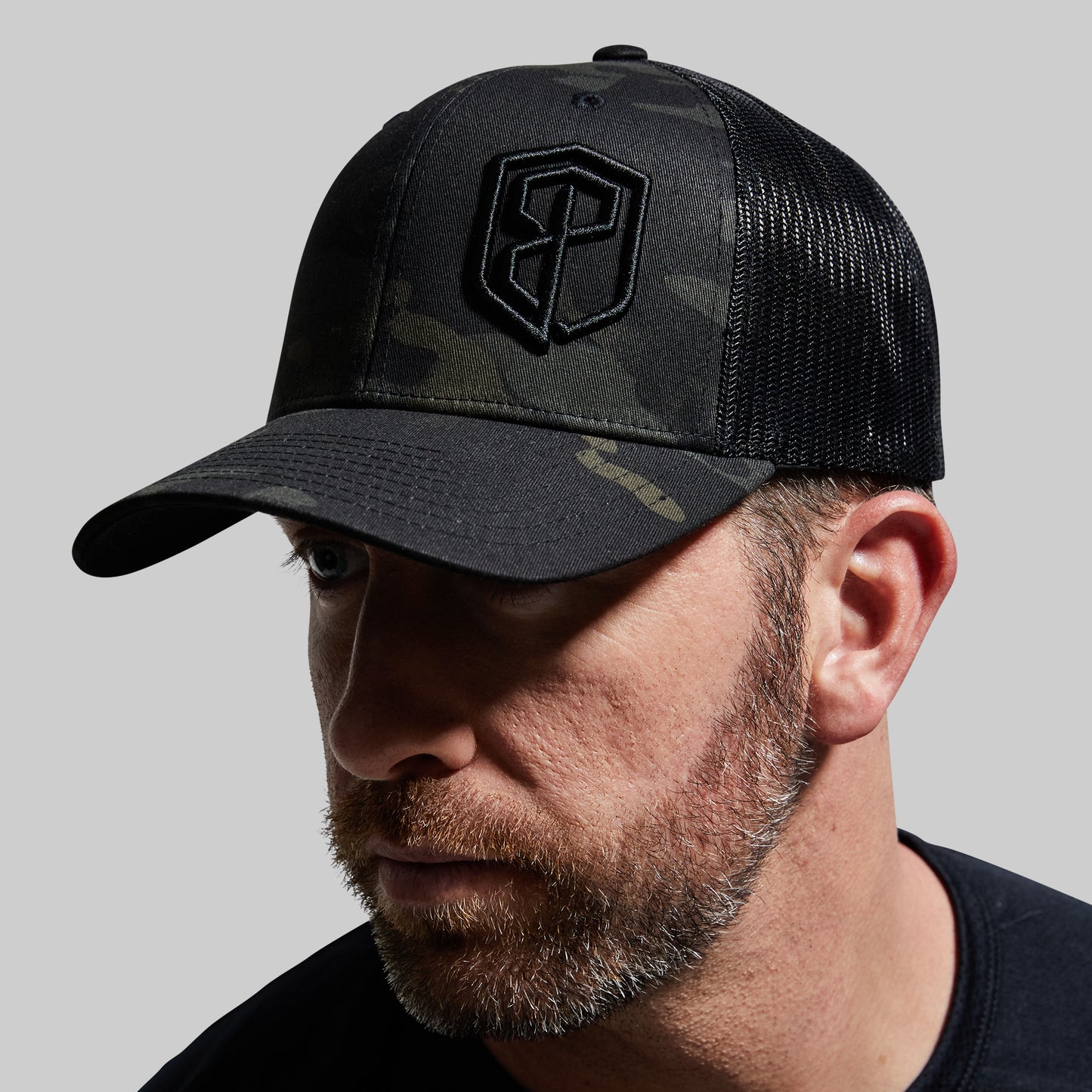 A person wearing a black multicam trucker hat with a camouflage pattern and a logo on the front.