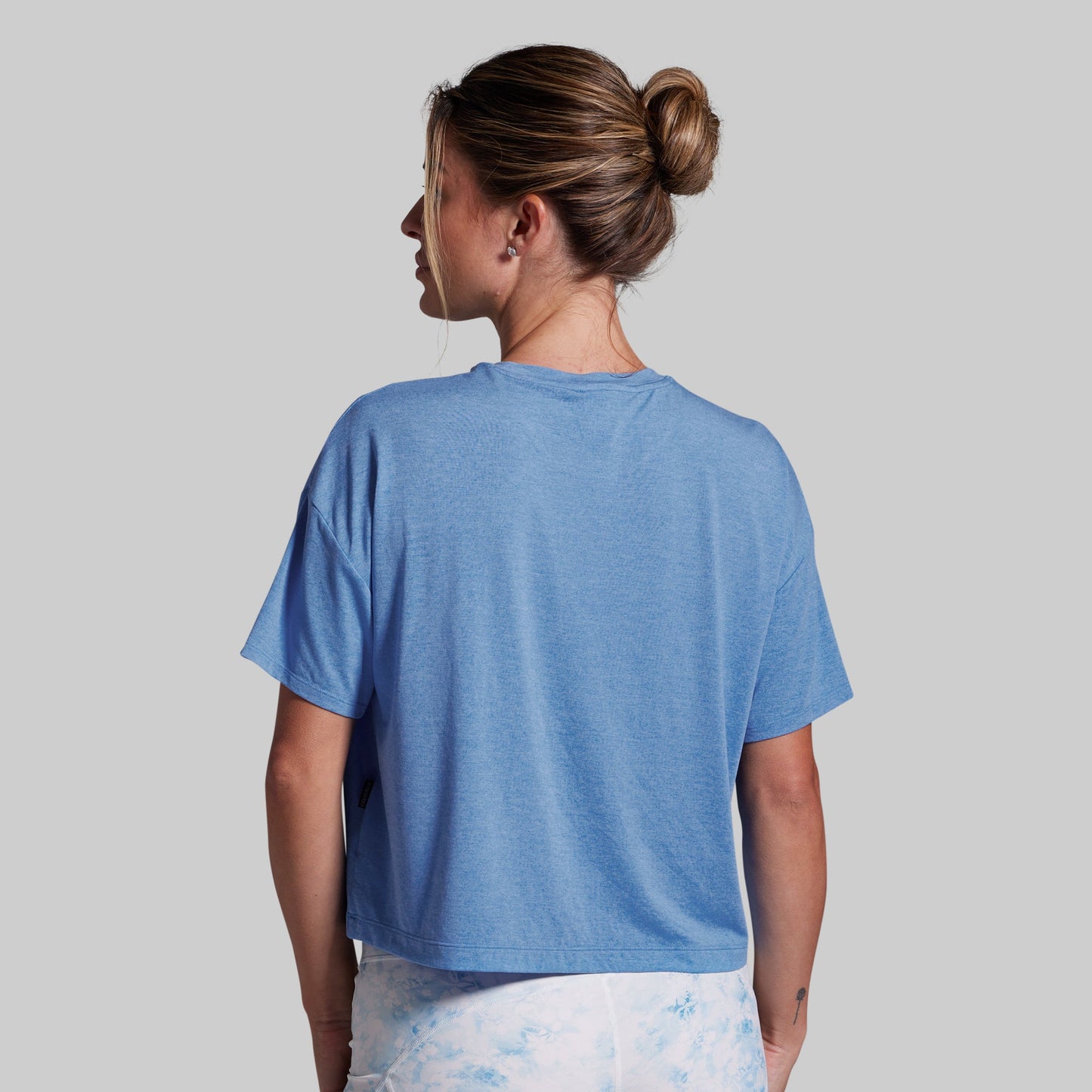 A woman viewed from the back wearing a heathered light blue short-sleeve crop top and white tie-dye pants, set against a solid light gray background.
