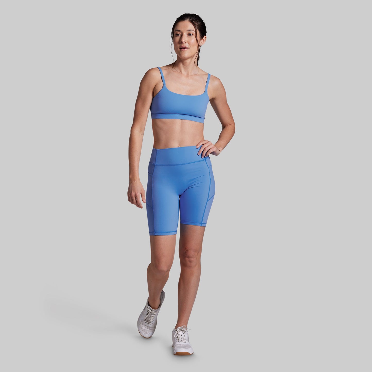 A woman modeling a matching athletic set consisting of a light blue sports bra and high-waisted biker shorts, paired with white sneakers, against a solid gray studio background.