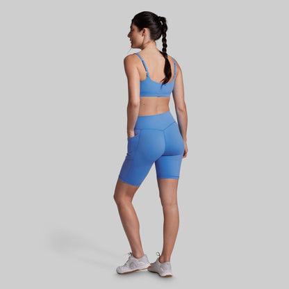 A woman wearing a matching athletic set consisting of a light blue sports bra and high-waisted biker shorts, shown from a rear three-quarter angle against a solid gray background.