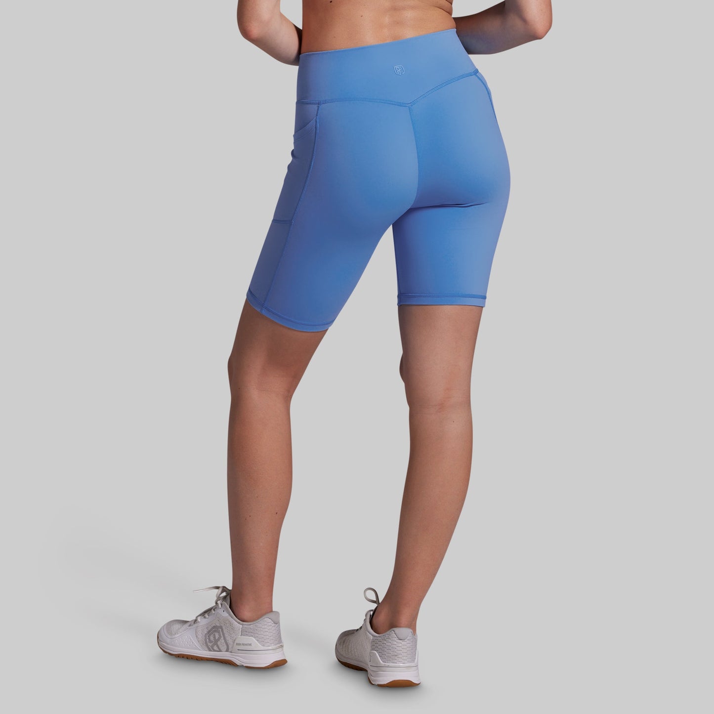 A model from the back wearing blue high-waisted biker shorts and light gray athletic sneakers, set against a solid light gray background.