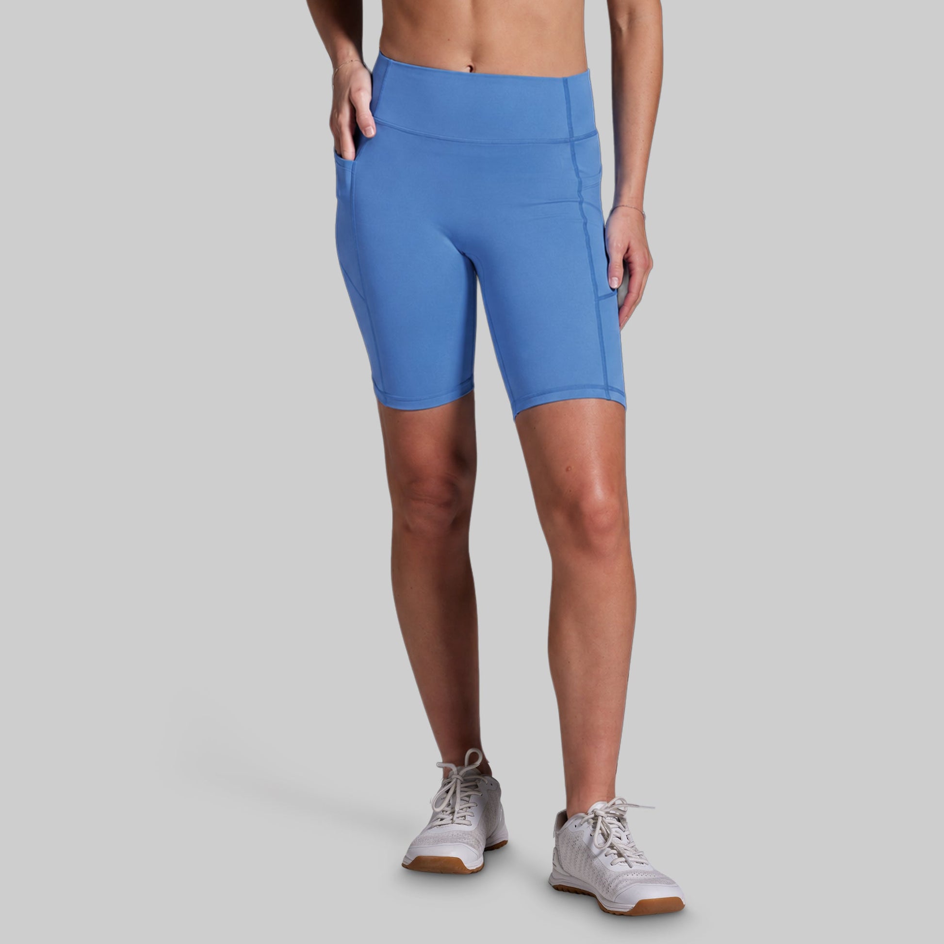 A model wearing bright blue athletic biker shorts with side pockets, paired with white sneakers, shown from the waist down against a solid gray background.