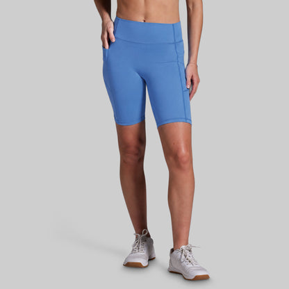 A model wearing bright blue athletic biker shorts with side pockets, paired with white sneakers, shown from the waist down against a solid gray background.
