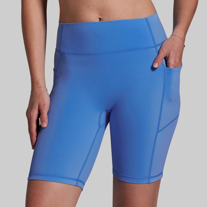 A close-up of a model wearing bright blue high-waisted biker shorts with side pockets, shown from the waist to mid-thigh against a light gray studio background.