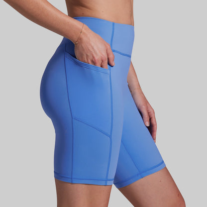 A close-up of a model wearing bright blue high-waisted athletic biker shorts with side pockets, shown from the waist to mid-thigh against a plain light gray background.