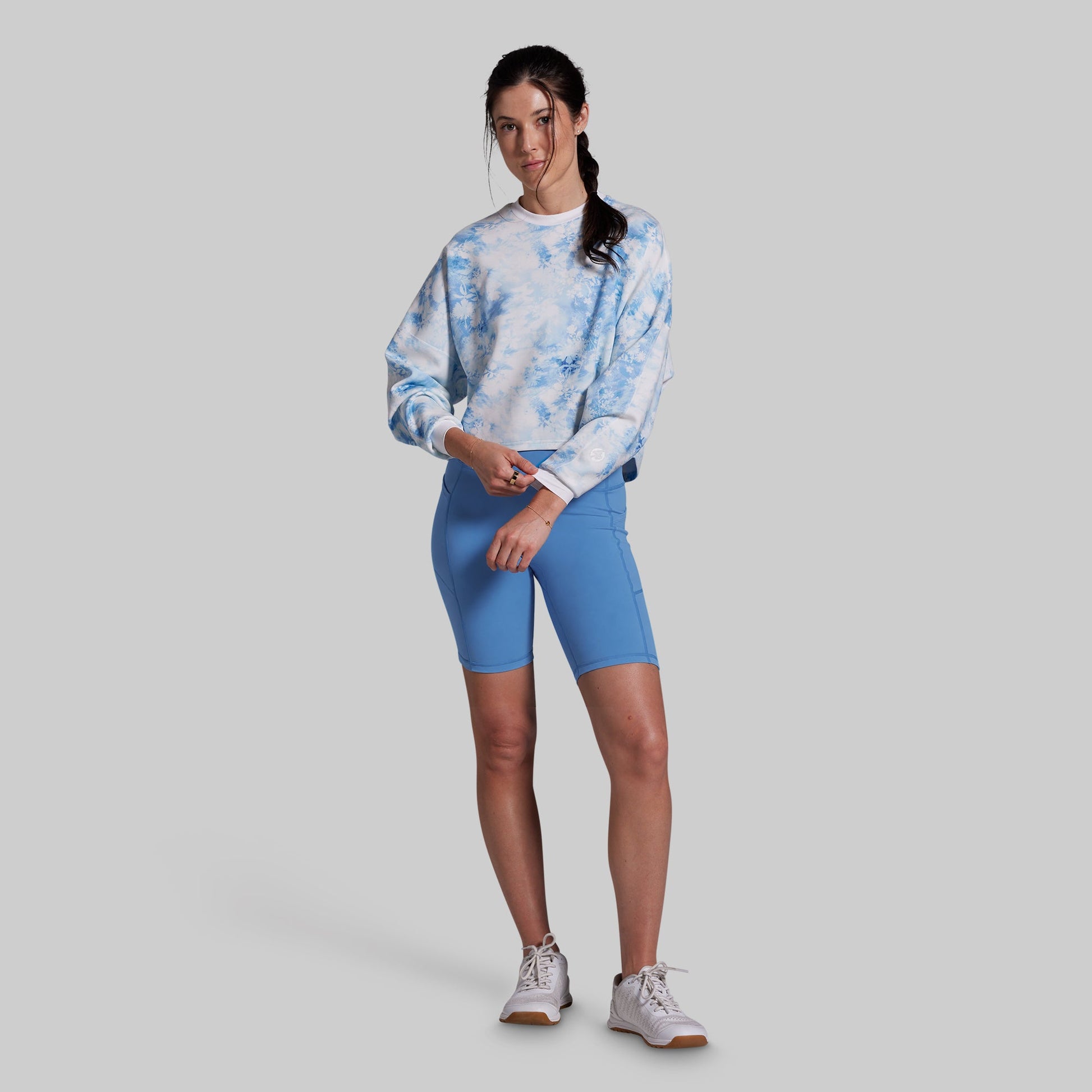 A woman modeling a light blue and white tie-dye sweatshirt paired with bright blue fitted biker shorts and white sneakers, standing against a solid gray studio background.