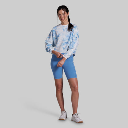 A woman modeling a light blue and white tie-dye sweatshirt paired with bright blue fitted biker shorts and white sneakers, standing against a solid gray studio background.