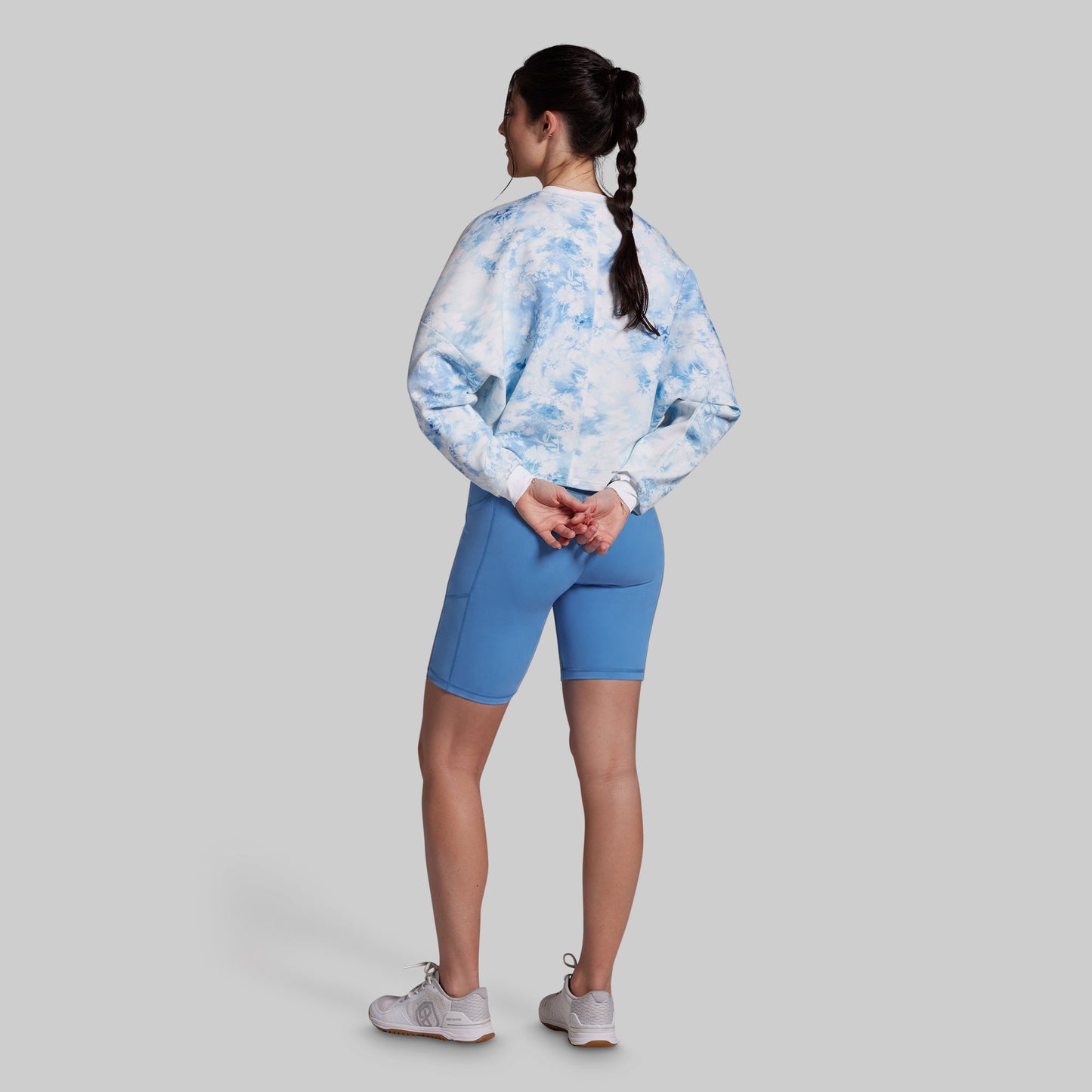 A woman shown from the back wearing a light blue and white tie-dye long-sleeve top, blue fitted biker shorts, and white sneakers, standing against a solid gray background.