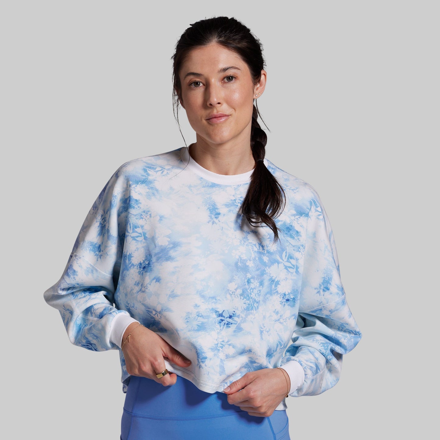 A woman modeling a light blue and white tie-dye crewneck sweatshirt, paired with blue pants, against a solid light gray background.